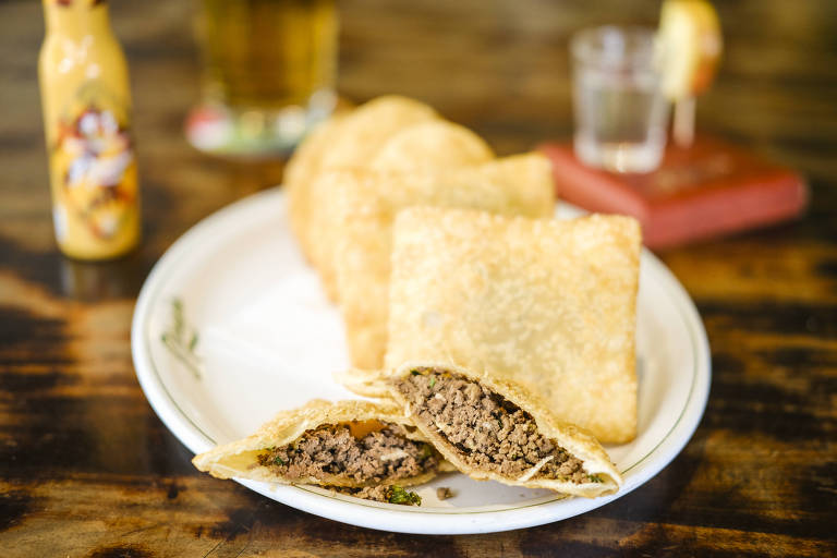 Um prato com pastéis de carne fritos, sendo um deles cortado ao meio, revelando o recheio de carne moída. Ao fundo, há mais pastéis e uma garrafa de molho de pimenta, além de um copo com bebida e uma fatia de limão.