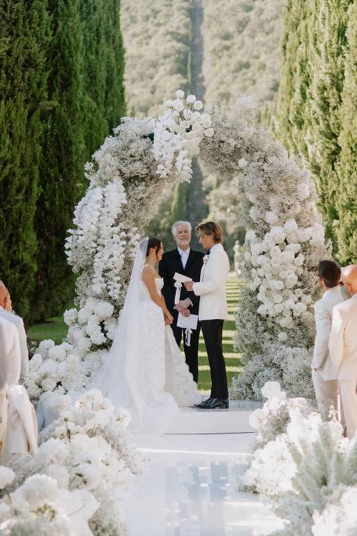 A imagem mostra uma cerimônia de casamento ao ar livre, com um arco floral decorado com flores brancas e folhagens