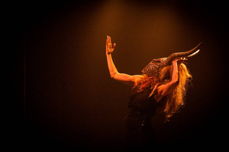 A imagem mostra uma performer em um palco iluminado com luzes quentes. Ela está usando uma máscara com chifres e tem cabelos longos e soltos. A performer está em uma pose expressiva, com um braço levantado e a cabeça inclinada para trás, criando uma atmosfera dramática.