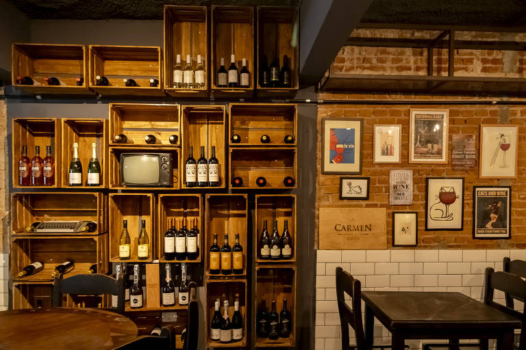 A imagem mostra o interior de um bar com prateleiras de madeira em forma de caixas, contendo garrafas de vinho. Há uma televisão antiga em uma das prateleiras. A parede à direita é decorada com várias molduras e quadros. O ambiente é acolhedor, com mesas e cadeiras de madeira visíveis.