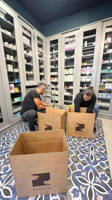 A imagem mostra duas pessoas organizando produtos em uma farmácia. Elas estão agachadas, colocando itens em caixas de papelão. Ao fundo, há prateleiras com diversos medicamentos e produtos de saúde. O chão é coberto por um tapete com um padrão decorativo. As caixas têm o logotipo 'BARAKA'.