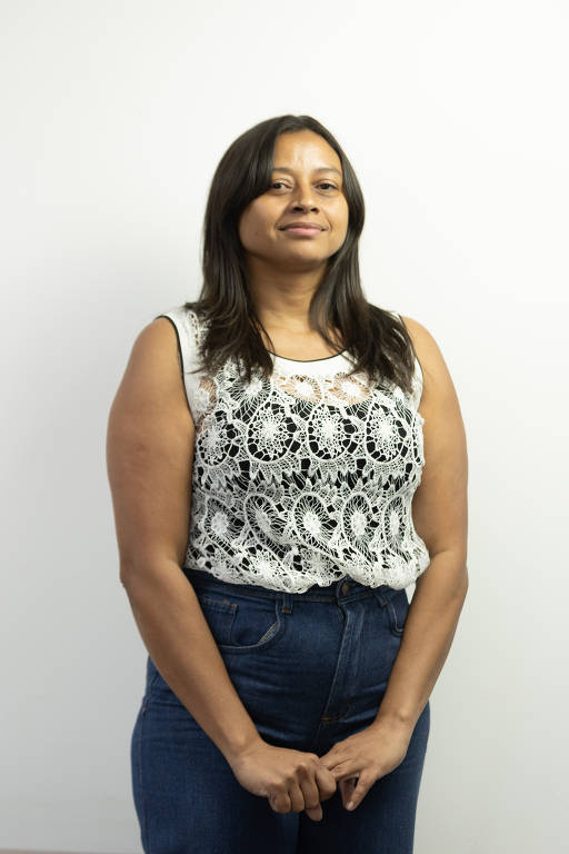 A imagem mostra uma mulher em pé, com um fundo branco. Ela usa uma blusa de renda branca com detalhes em preto e jeans escuros. A mulher está com as mãos unidas na frente do corpo e tem uma expressão neutra.