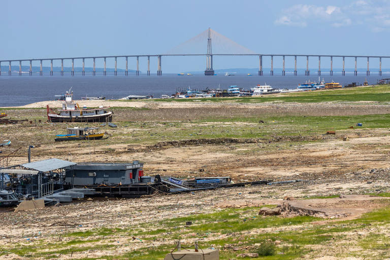 A imagem mostra uma paisagem costeira com uma ponte longa e elevada ao fundo, cruzando um corpo de água. No primeiro plano, há uma área de terra seca com vegetação esparsa e alguns barcos e estruturas de metal. O céu está claro com algumas nuvens.