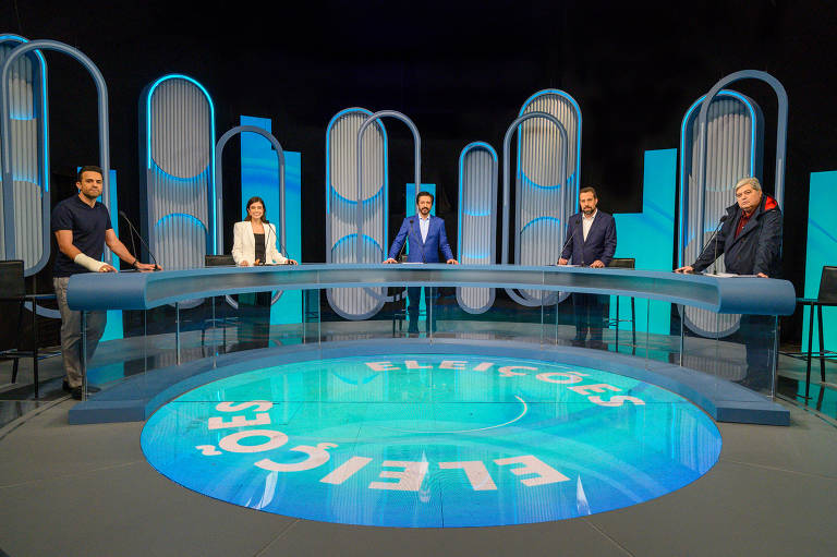 A imagem mostra um estúdio de televisão com cinco pessoas em um painel de debate. Elas estão posicionadas ao redor de uma mesa circular. O fundo é composto por estruturas verticais em tons de azul e branco, e o chão tem a palavra 'Eleições' em destaque. As pessoas estão vestidas de maneira formal, com três homens e duas mulheres visíveis.