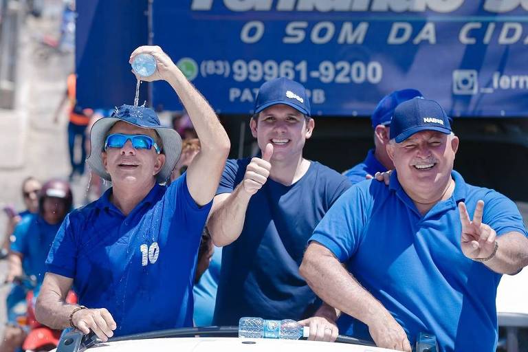 A imagem mostra três pessoas em um carro durante um evento ao ar livre. Duas delas estão vestidas com camisetas azuis e bonés， uma delas levantando uma garrafa de água， enquanto a outra faz um gesto de positivo com o polegar. A terceira pessoa， também vestida de azul， faz um sinal de paz com a mão. Ao fundo， há uma faixa com o nome 039;Fernando Silva039; e informações de contato.