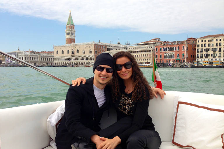 Pavel Durov e Irina Bolgar estão sentados em um barco, sorrindo e posando para a foto. Eles estão vestidos de forma casual, com Pavel Durov, que é um homem usando um boné e óculos escuros, e Irina Bulgar, que é uma mulher com óculos escuros e uma blusa escura. Ao fundo, é possível ver a arquitetura de Veneza, incluindo um campanário e edifícios coloridos, com água ao redor.