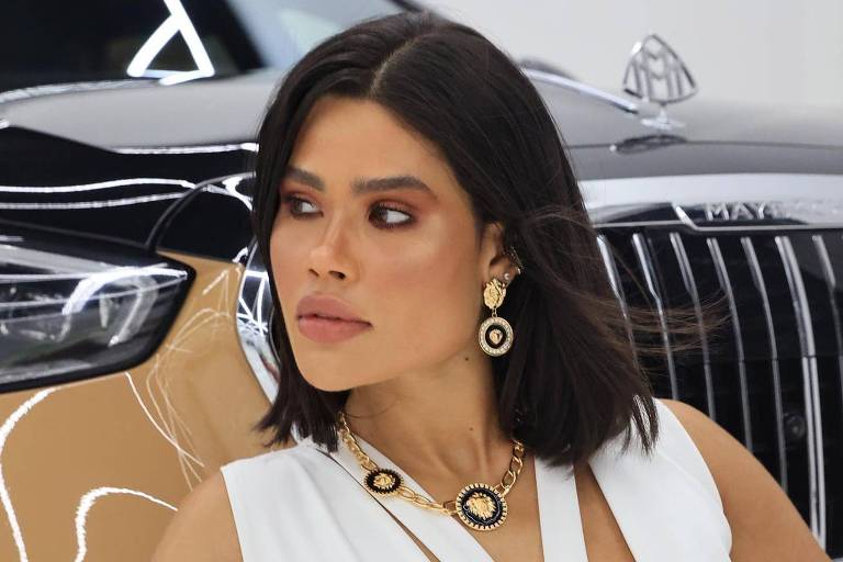 Uma mulher com cabelo liso e escuro, usando um vestido branco com recortes, está posando em frente a um carro de luxo