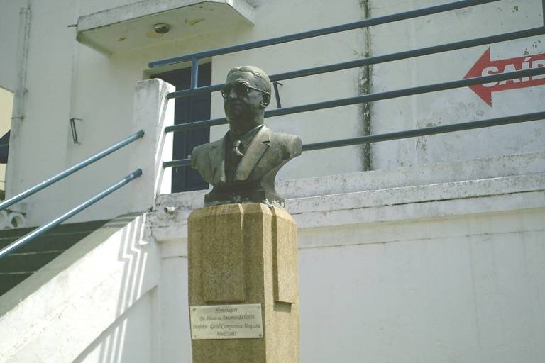 A imagem mostra um busto de uma figura histórica em um pedestal de madeira. O busto está localizado em uma área externa, próximo a uma escada e uma parede branca. Ao fundo, há uma estrutura com corrimãos e uma seta vermelha indicando direção