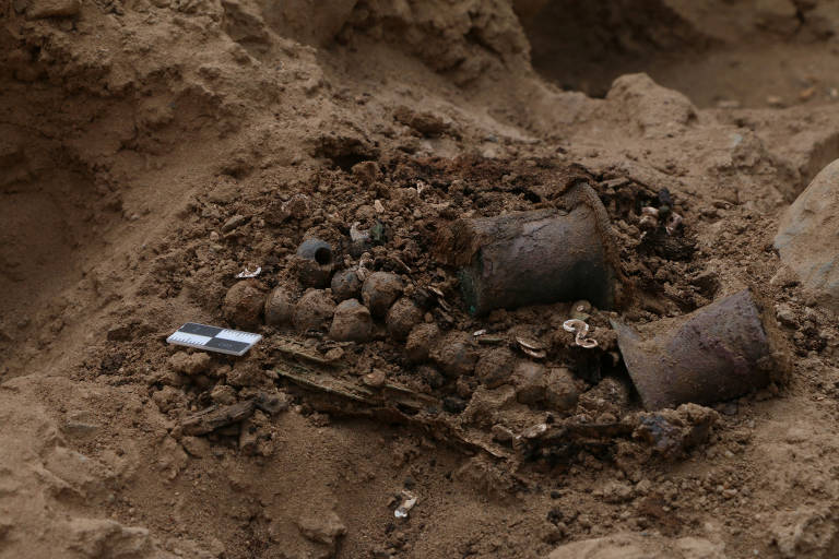 A imagem mostra uma área de escavação com um monte de terra e vários objetos enterrados. Entre os itens visíveis, há dois recipientes de metal, esferas de metal e fragmentos de material orgânico. Um pequeno objeto retangular, possivelmente uma régua de medição, está ao lado dos itens. O solo ao redor é arenoso e irregular.