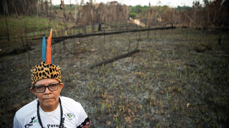 Um homem indígena está em pé em um terreno queimado, com árvores carbonizadas ao fundo. Ele usa um chapéu com penas coloridas e uma camiseta branca. O solo ao redor é escuro e seco, com algumas plantas verdes começando a brotar.