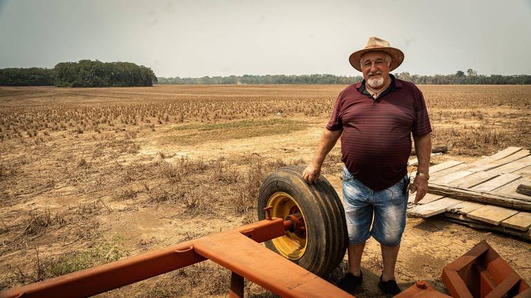 Um homem idoso, usando um chapéu e uma camisa roxa, está posando ao lado de um pneu em um campo seco. Ele usa shorts jeans e está em uma área rural com um solo árido ao fundo. A paisagem é ampla, com algumas árvores distantes e um céu claro.