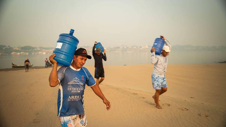 A imagem mostra três homens caminhando na areia de uma praia, cada um carregando um grande galão azul de água sobre os ombros. O homem mais próximo está usando uma camiseta azul com detalhes brancos e shorts claros. Os outros dois homens estão mais ao fundo, um deles vestindo uma camiseta de manga longa e o outro com uma camiseta de manga curta. O ambiente é ensolarado e há fumaça no ar, com uma cidade visível ao fundo.