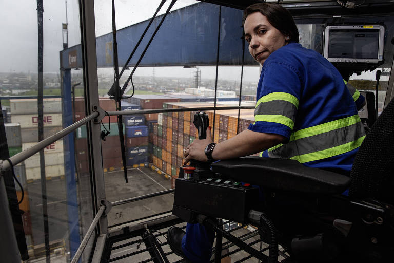 A imagem mostra uma mulher sentada em um guindaste, olhando para fora da cabine. Ela está vestindo um uniforme azul com faixas refletivas verdes. Ao fundo, há contêineres empilhados em um terminal, e o céu parece nublado.