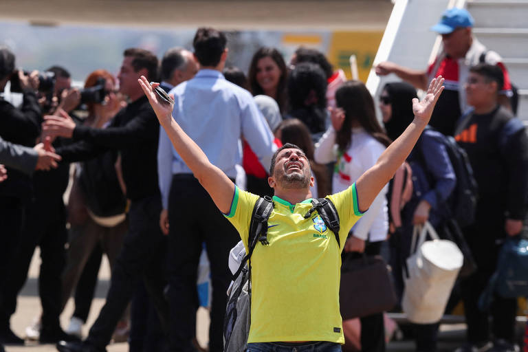 Um homem vestindo uma camiseta amarela com detalhes verdes, representando o Brasil, está de braços abertos em um gesto de celebração ou alegria. Ele está em frente a um grupo de pessoas que parecem estar recebendo alguém, com algumas segurando celulares e outras bolsas. O ambiente parece ser um aeroporto, com uma escada de embarque ao fundo.