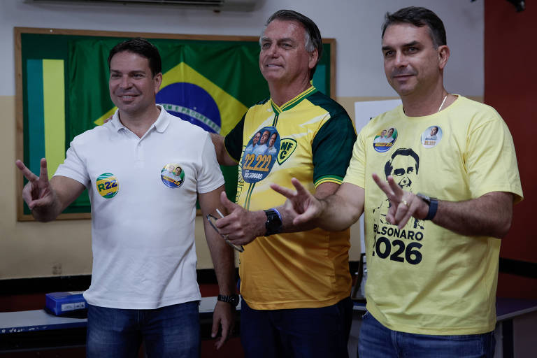 Três homens posam para a foto em um ambiente interno. Jair bolsonaro, o homem do meio, usa uma camisa verde e amarela, enquanto os outros dois vestem camisetas brancas e amarelas. Todos fazem o sinal de 'V' com as mãos, simbolizando apoio. Ao fundo, há uma bandeira do Brasil.