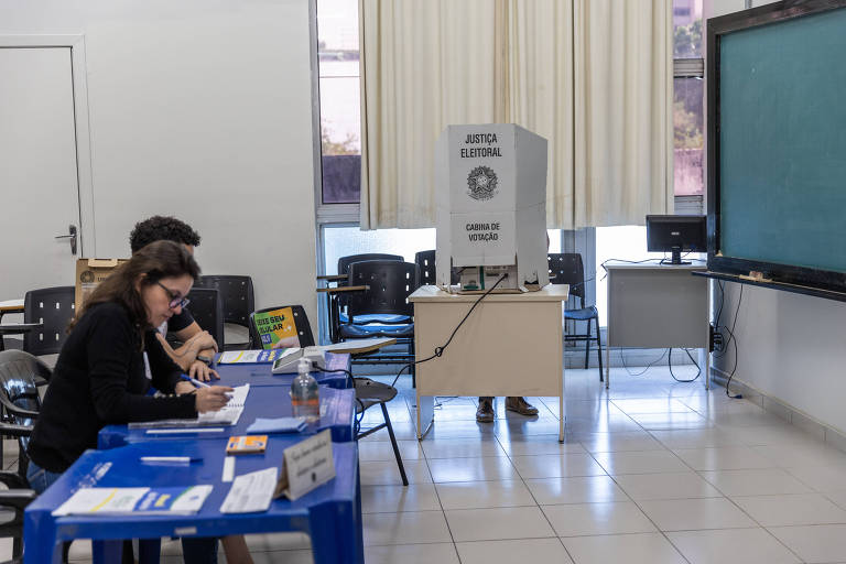 A imagem mostra uma sala de aula com duas mesas azuis. À esquerda, uma mulher está escrevendo em um caderno, enquanto um homem ao lado dela também está concentrado em sua atividade. No fundo, há um quadro branco e um equipamento de som ou projetor em uma mesa. As janelas estão cobertas com cortinas claras, permitindo a entrada de luz natural.
