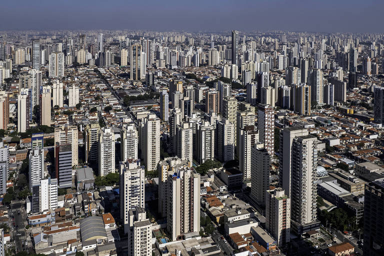 Imagem mostrando uma vasta área urbana com muitos edifícios altos e de diferentes tamanhos. A cidade é caracterizada por uma densa concentração de prédios， com ruas visíveis entre eles. O céu está claro， e a luz do sol ilumina a cena， destacando a arquitetura das construções.