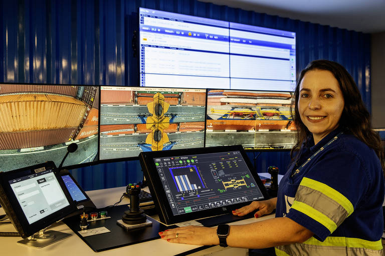 A imagem mostra uma mulher sentada em frente a um conjunto de monitores em um centro de controle. Ela está usando um uniforme azul com detalhes amarelos e sorri para a câmera. Os monitores exibem gráficos e dados， enquanto um tablet está posicionado à esquerda dela. O ambiente é moderno e organizado， com paredes azuis ao fundo.
