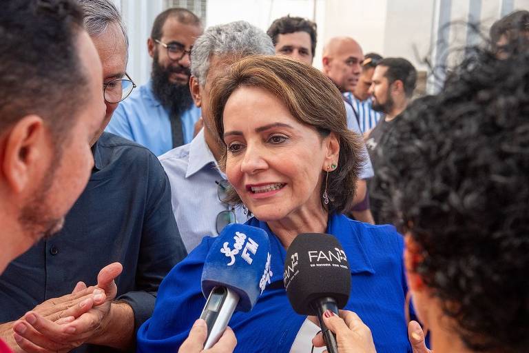 A imagem mostra uma mulher com cabelo castanho claro e liso, usando uma blusa azul, sendo entrevistada por repórteres. Ela está cercada por um grupo de pessoas, algumas com barba e óculos, que parecem estar ouvindo atentamente. Microfones de diferentes emissoras estão apontados para ela, enquanto ela sorri e fala.