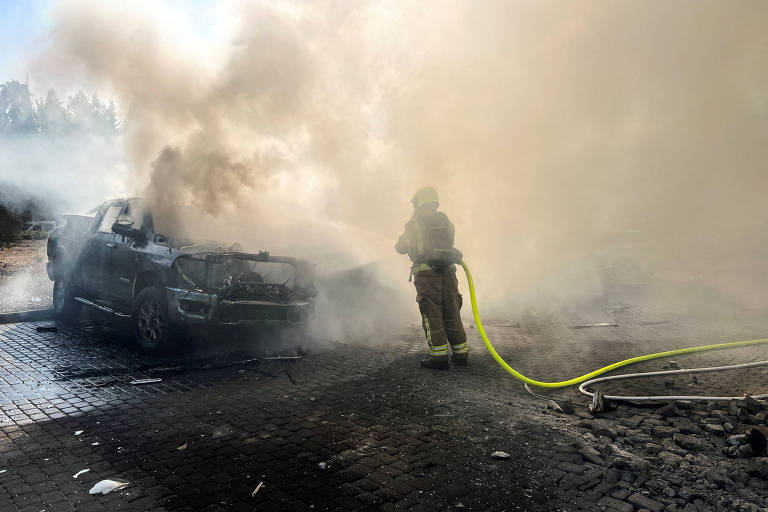 A imagem mostra um bombeiro em um local com muita fumaça, utilizando uma mangueira para combater um incêndio em um carro que está parcialmente queimado. O ambiente é coberto por uma densa nuvem de fumaça, dificultando a visibilidade. O carro está em uma superfície pavimentada.