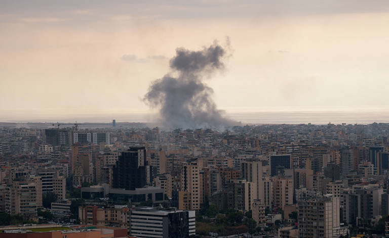 A imagem mostra uma vista panorâmica de uma cidade, com muitos edifícios e estruturas urbanas. No fundo, há uma nuvem de fumaça se elevando, indicando uma possível ocorrência de incêndio ou explosão. O céu está nublado, e o mar é visível ao longe.