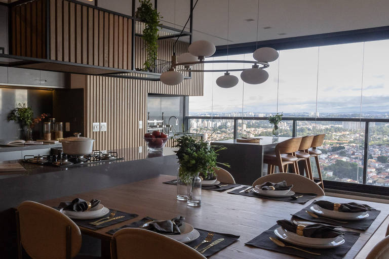 A imagem mostra uma cozinha moderna e bem iluminada, com uma ilha central e uma mesa de jantar. A cozinha possui armários escuros, uma geladeira prateada e um fogão a gás. Acima da ilha, há prateleiras com plantas e utensílios. A mesa de jantar está posta com pratos brancos e guardanapos cinzas, e há um vaso com flores. Ao fundo, uma grande janela oferece uma vista panorâmica da cidade.