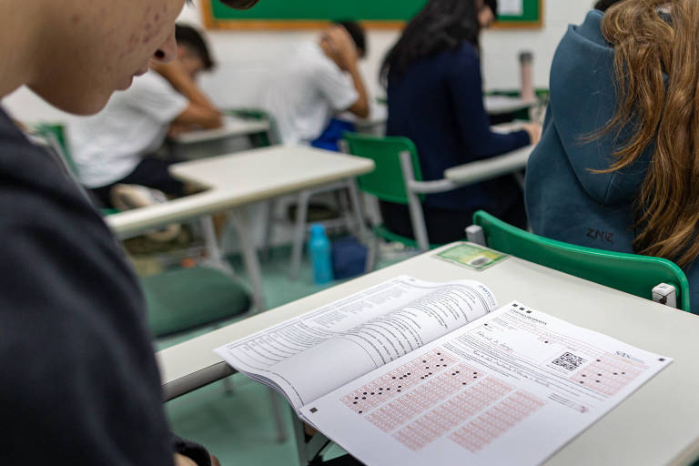 A imagem mostra um estudante sentado em uma sala de aula， olhando para um caderno aberto sobre a mesa. Ao fundo， outros alunos estão concentrados em suas atividades. O caderno contém gráficos e informações， incluindo um código QR. As mesas são de cor clara e as cadeiras são verdes.