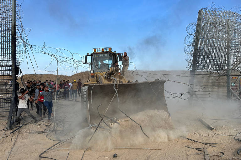 A imagem mostra uma máquina de construção, possivelmente uma retroescavadeira, derrubando uma barreira de arame farpado em um ambiente desértico. Há poeira levantada pela ação da máquina e um grupo de pessoas observando a cena ao fundo. O céu está claro e azul.