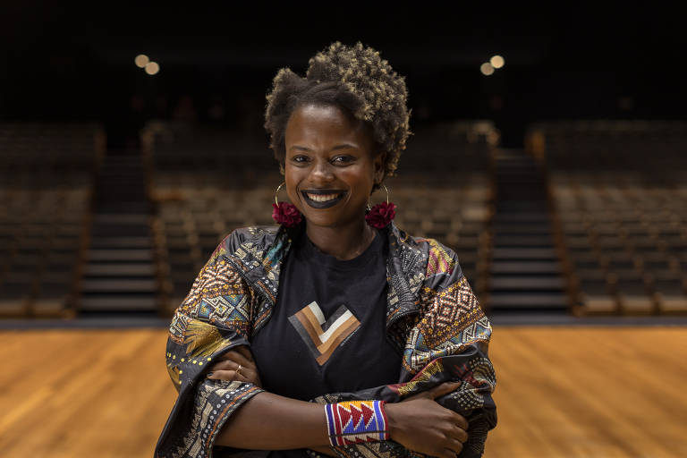 A imagem mostra uma mulher sorridente em um ambiente de teatro. Ela tem cabelo crespo e usa um batom escuro. Está vestida com uma camiseta preta e um casaco colorido com padrões. A mulher tem brincos grandes e flores na orelha, além de uma pulseira colorida no braço. Ao fundo, há assentos de teatro vazios.