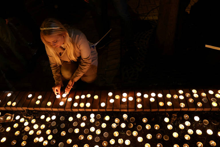 Uma mulher de cabelo loiro está agachada, acendendo uma vela em uma linha de velas acesas em um espaço escuro. As velas estão dispostas em uma superfície de tijolos, com muitas outras velas acesas ao redor, criando uma atmosfera de reflexão e homenagem.