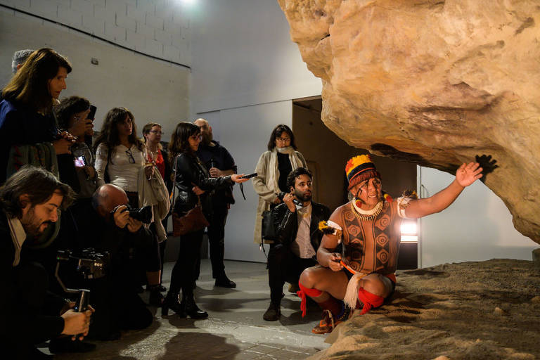 A imagem mostra um grupo de pessoas em um ambiente de museu. Um homem vestido com trajes tradicionais, incluindo um cocar colorido, está agachado e gesticulando em direção a uma rocha. Ele segura um objeto em uma das mãos. O público, composto por várias pessoas, está atento e alguns estão tirando fotos. O ambiente é iluminado e possui paredes brancas.