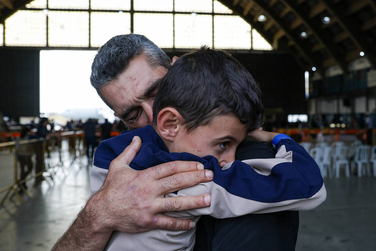 A imagem mostra um homem abraçando um menino em um ambiente amplo, possivelmente um galpão ou armazém. O homem está com o rosto próximo ao menino, que parece estar se aconchegando em seu peito. O espaço ao fundo é iluminado, com várias cadeiras brancas dispostas e uma grande janela ao fundo.