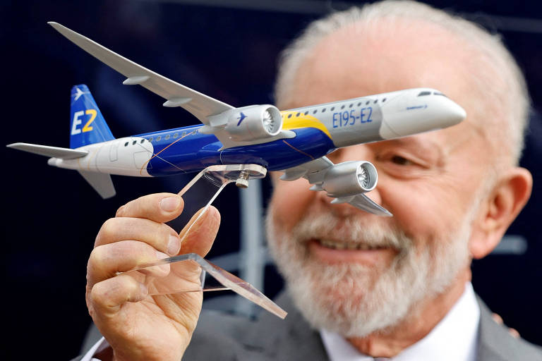 Um homem idoso com barba branca sorri enquanto segura um modelo de avião em miniatura. O avião é colorido, com a fuselagem em azul e amarelo, e a letra 'E2' visível na cauda. O fundo é desfocado.