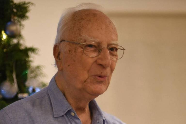 Um homem idoso com cabelo grisalho e óculos, usando uma camisa azul. Ele está em um ambiente interno, com uma árvore de Natal decorada ao fundo. O homem parece estar falando ou expressando uma ideia.