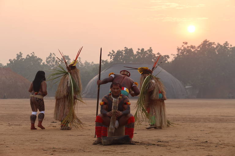 A imagem mostra um grupo de pessoas indígenas em um ambiente ao ar livre durante o amanhecer. Um homem está sentado em um tronco, segurando um objeto, enquanto outros estão em pé, alguns com adornos de palha e penas. O cenário é em um terreno de terra batida, com algumas habitações ao fundo e a luz suave do sol nascente.