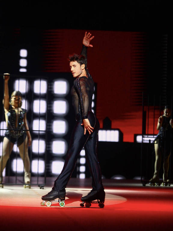 Um artista masculino está se apresentando em um palco iluminado, usando um traje justo e patins. Ele está em uma pose dinâmica, com um braço levantado e o corpo virado para o lado. Ao fundo, há uma tela com luzes e formas geométricas, e duas dançarinas estão visíveis atrás dele, uma delas em uma jaula. O cenário é predominantemente vermelho.