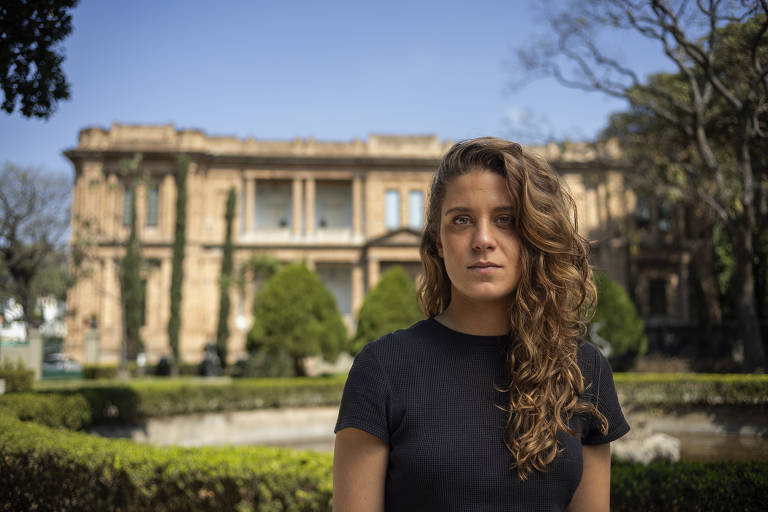 Uma mulher com cabelo cacheado e longo está em primeiro plano, vestindo uma camiseta preta. Ao fundo, há um parque com árvores e um edifício de tijolos visível. A cena é iluminada por luz natural, com um céu azul e algumas nuvens. A mulher parece estar olhando diretamente para a câmera.