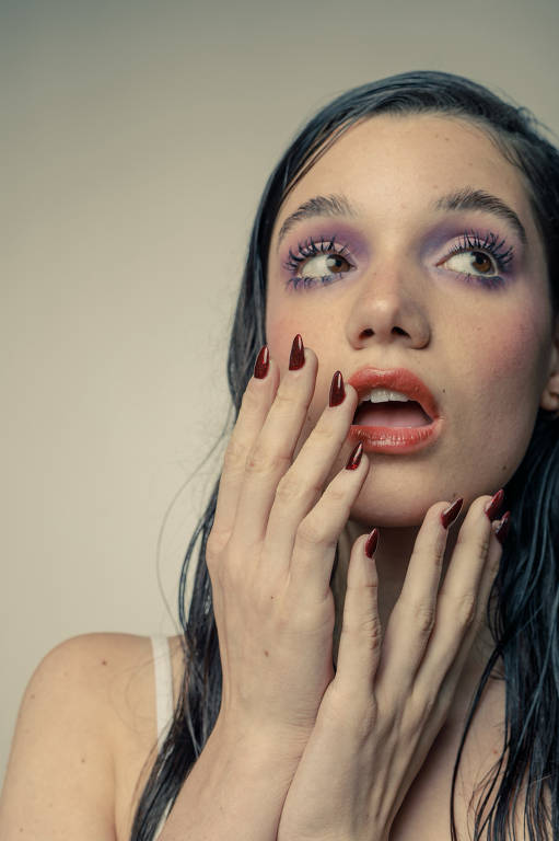 A imagem mostra uma mulher com cabelo molhado e maquiagem artística. Ela tem olhos destacados com sombra roxa e cílios longos. Seus lábios estão pintados de vermelho e suas unhas são longas e de cor vermelha brilhante. A mulher está com as mãos em seu rosto, expressando surpresa ou admiração, e o fundo é de uma cor neutra.