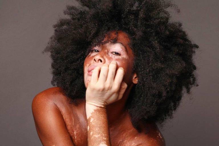 A imagem mostra uma mulher com cabelo afro volumoso, sentada em uma pose expressiva. Ela está com a mão cobrindo parcialmente o rosto e tem tatuagens visíveis nos braços. A pele dela é de um tom bronzeado e o fundo é neutro, destacando a figura.