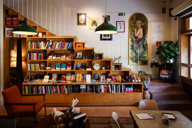 A imagem mostra o interior de uma livraria com uma estante de livros em madeira, que ocupa uma parede e se estende até um nível superior. Há uma poltrona laranja à esquerda e mesas de madeira na parte inferior. O ambiente é iluminado por lâmpadas pendentes verdes e possui uma planta ao fundo, além de uma pintura decorativa na parede. O espaço é acolhedor e convidativo.