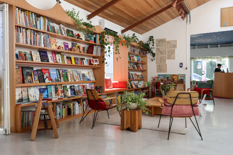 A imagem mostra o interior de uma biblioteca com uma estante de madeira cheia de livros. Há plantas decorativas ao redor e cadeiras vermelhas confortáveis. A iluminação é suave, com uma janela que deixa entrar luz natural. No fundo, pode-se ver uma área de recepção.