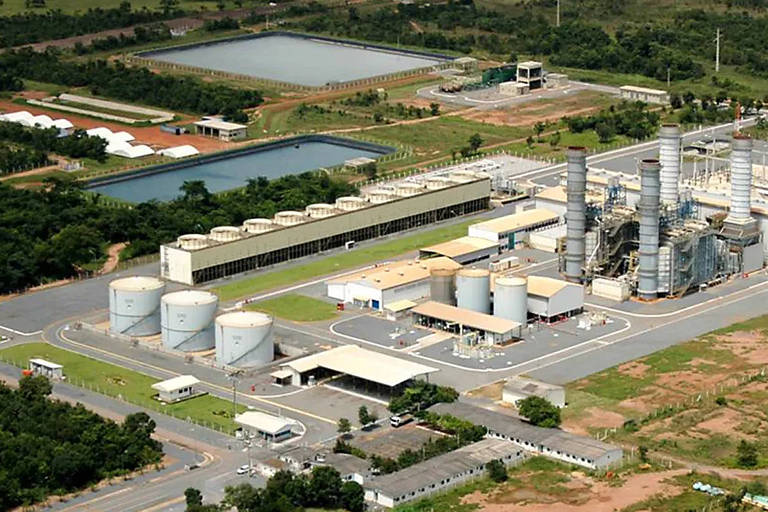 A imagem mostra uma vista aérea de uma instalação industrial. Há tanques grandes em primeiro plano， uma estrutura de fábrica com chaminés e silos. Ao fundo， há áreas de vegetação e tanques de água. O ambiente é predominantemente verde， com áreas pavimentadas e construções.