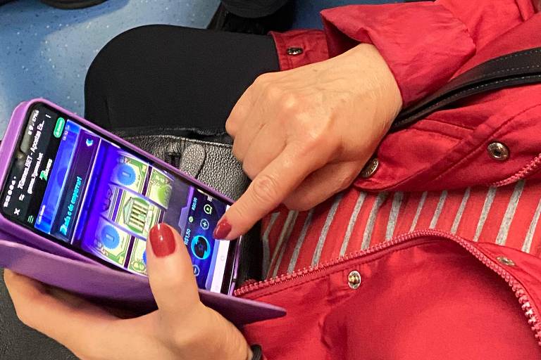 Uma mão feminina com unhas pintadas de vermelho está tocando a tela de um celular que está em uma capa roxa. O celular exibe um aplicativo com gráficos coloridos. A pessoa está vestindo uma jaqueta vermelha e há uma blusa listrada visível. O fundo mostra o piso azul de um transporte público e parte de outras pessoas ao redor.
