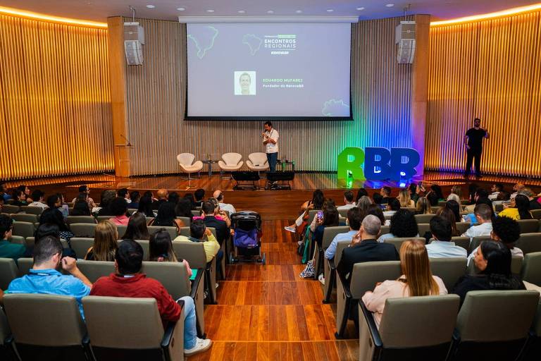 Pessoas sentadas em auditório com palestrante no palco falando ao microfone