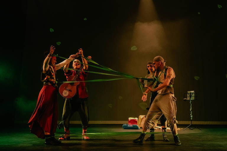 A imagem mostra quatro artistas em um palco durante uma apresentação teatral. Dois deles estão segurando fitas verdes, enquanto um deles toca um violão. Os artistas estão vestidos de forma colorida e expressiva, com um fundo escuro e iluminação suave. No chão, há alguns objetos, como uma caixa e um suporte para partituras. Folhas parecem estar caindo ao redor.