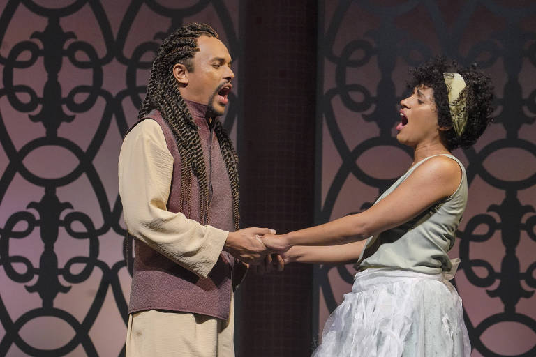 A imagem mostra dois cantores em uma apresentação teatral. O homem à esquerda, com cabelo trançado e vestindo uma túnica clara e um colete escuro, está segurando as mãos da mulher à direita, que usa uma blusa verde e uma saia branca. Ambos estão cantando com expressões intensas, em um cenário com um fundo decorativo que apresenta padrões em preto e tons de rosa.