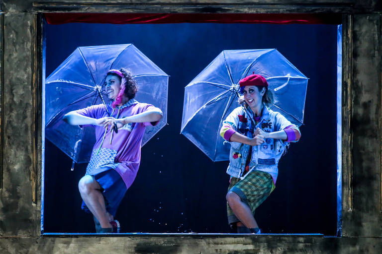 Duas performers estão em um palco, cada uma segurando um guarda-chuva transparente. Elas estão em uma moldura que parece uma janela, com um fundo escuro. A artista à esquerda usa uma camiseta rosa e shorts, enquanto a da direita veste um colete jeans decorado e uma boina vermelha. Ambas estão em poses dinâmicas, sorrindo e aparentando estar em uma performance animada.
