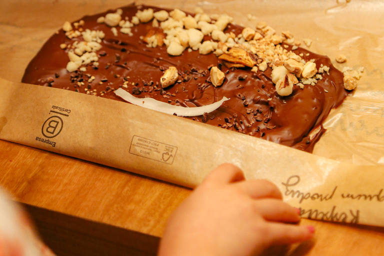 A imagem mostra uma porção de chocolate derretido coberta com pedaços de nozes e granola, disposta sobre um papel manteiga. Uma mão infantil segura a borda do papel, enquanto o fundo revela uma mesa de madeira. Há anotações visíveis no papel, incluindo palavras como 'Umaro' e 'Kekanto'.