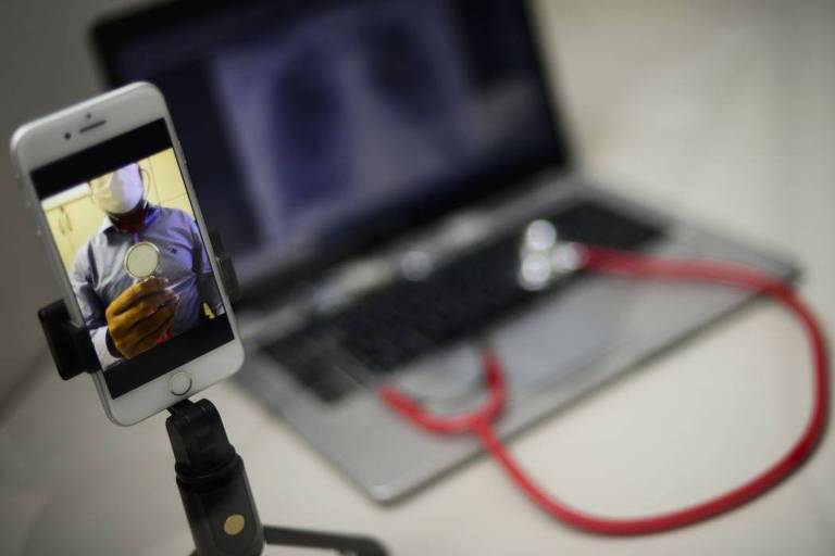 No primeiro plano da imagem， há um celular em um suporte com a imagem de um médico em teleatendimento. No segundo， um notebook com uma estetoscópio vermelho.  