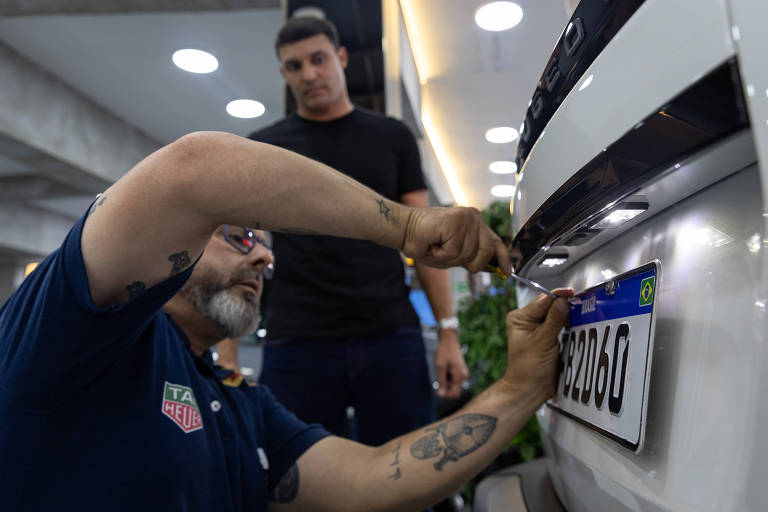 Homem com barba e óculos, usando uma camisa azul, está instalando uma placa de veículo na parte traseira de um carro. Ele está usando uma chave de fenda e parece concentrado na tarefa. Ao fundo, um jovem de camiseta preta observa o processo. O ambiente é bem iluminado, com paredes de concreto e elementos decorativos